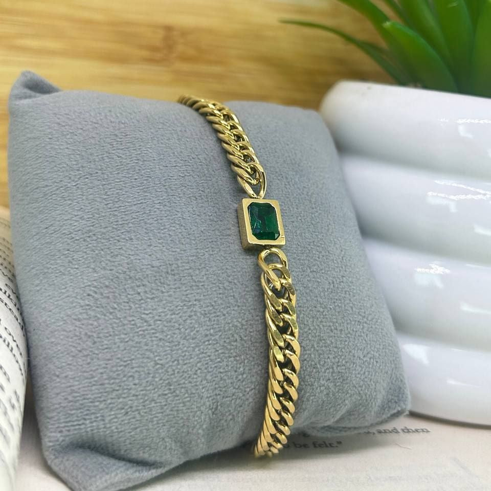 Gold bracelet with a green stone on a gray pillow with a plant in the background