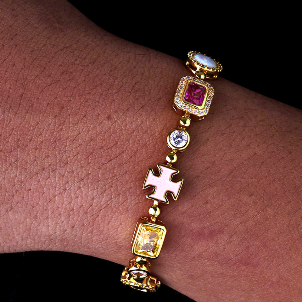 A gold-plated charm bracelet with multiple links, including a Maltese cross, a yellow gemstone, a pink gemstone, and a white gemstone, shown worn on a person's wrist against a black background.