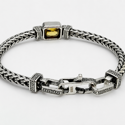 A silver-toned braided chain bracelet with a rectangular yellow gemstone centerpiece and decorative Greek key link details, shown on a light gray background.