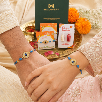 A close-up of a person's hands wearing two matching blue and gold Rakhi bracelets, with product packaging and a green Mr. Dapper's box placed on a decorative tray in the background.