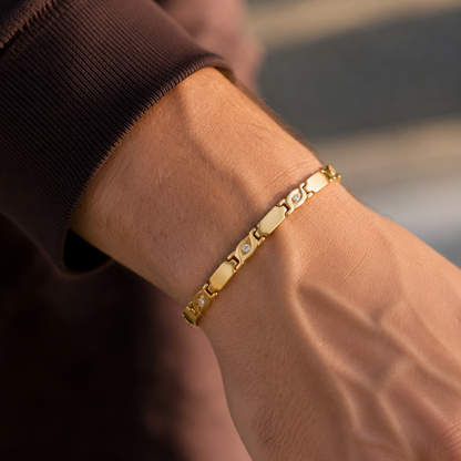 A close-up of a gold link bracelet with oval and heart-shaped links, worn on a person's wrist. The person is wearing a dark brown ribbed long-sleeve top, and the background is a softly blurred outdoor street scene.