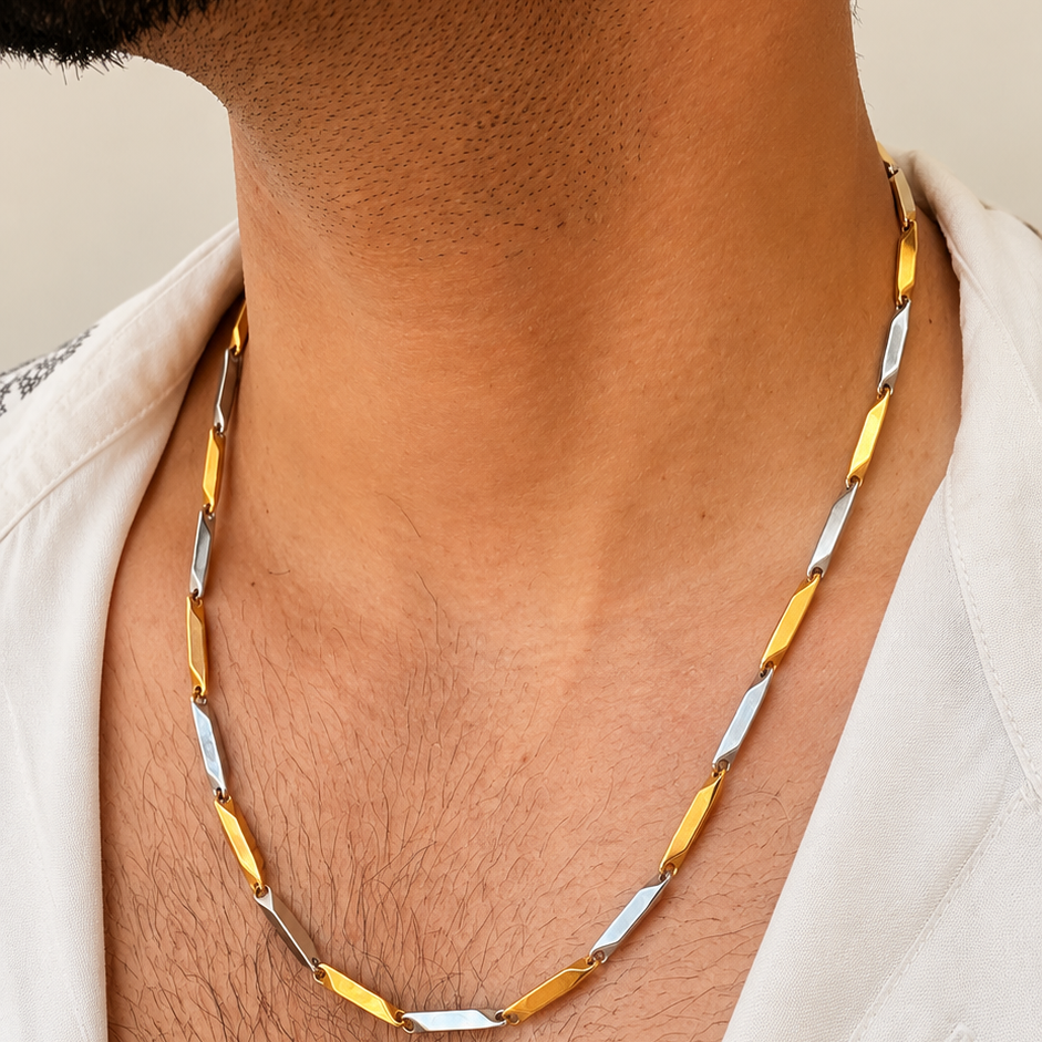 A close-up of a man's neck and chest wearing a twisted chain necklace with alternating gold and silver-toned links, styled with a white open-collar shirt.