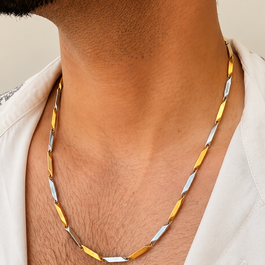 A close-up of a man's neck and chest wearing a twisted chain necklace with alternating gold and silver-toned links, styled with a white open-collar shirt.