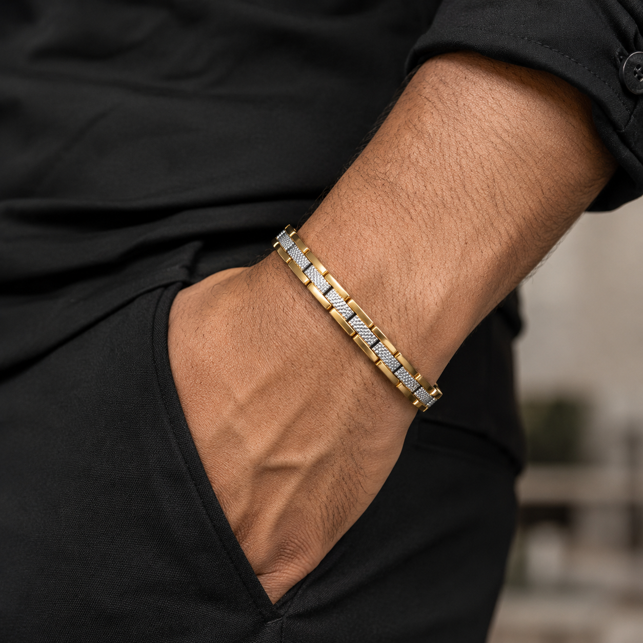 A close-up of a man's forearm wearing a gold and silver chain-link bracelet, dressed in a black shirt and black pants.