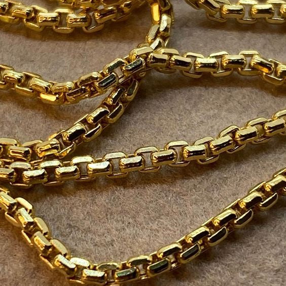 A close-up of a shiny gold chain necklace with a curb-style link pattern, draped across a textured beige surface.