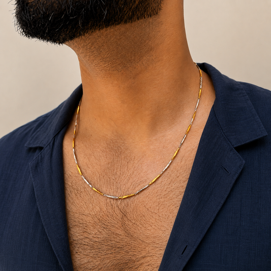 A close-up of a man wearing a delicate two-tone chain necklace with gold and silver beads, styled with a navy blue shirt.