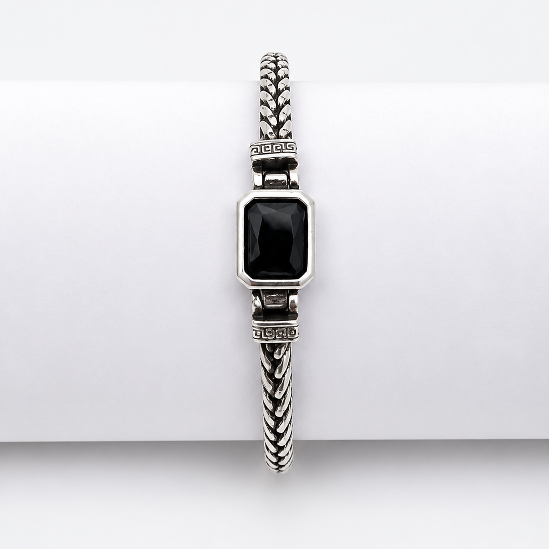 A silver-toned braided cable bracelet with a rectangular black inlay, displayed on a white cylindrical stand against a light gray background.