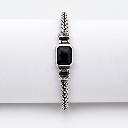A silver-toned braided cable bracelet with a rectangular black inlay, displayed on a white cylindrical stand against a light gray background.