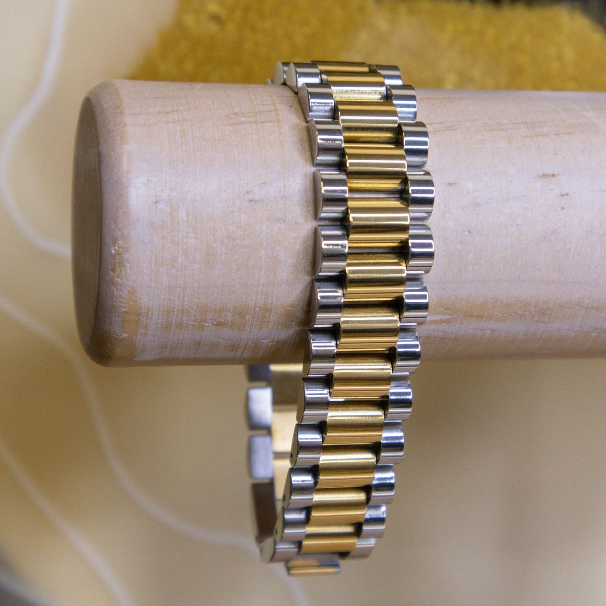 A two-tone metal link bracelet with alternating gold and silver cylindrical links, displayed on a light wood cylindrical stand against a beige background with a yellow textured area.