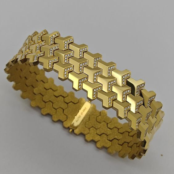 A gold-tone geometric link bracelet with a top row of interlocking cube-like links set with small clear stones and a lower row of textured hexagonal links, shown on a plain light gray background.