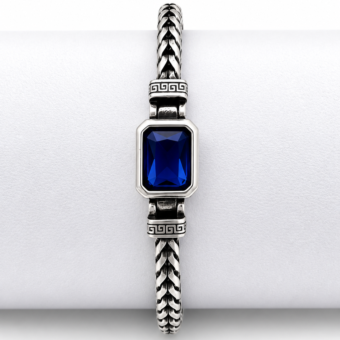 A silver-toned braided cable bracelet with a rectangular deep blue gemstone centerpiece, displayed on a white cylindrical ring.