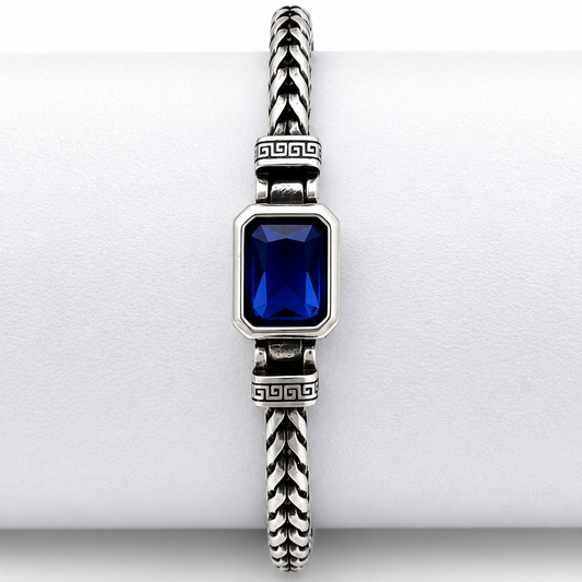 A silver-toned braided cable bracelet with a rectangular deep blue gemstone centerpiece, displayed on a white cylindrical ring.