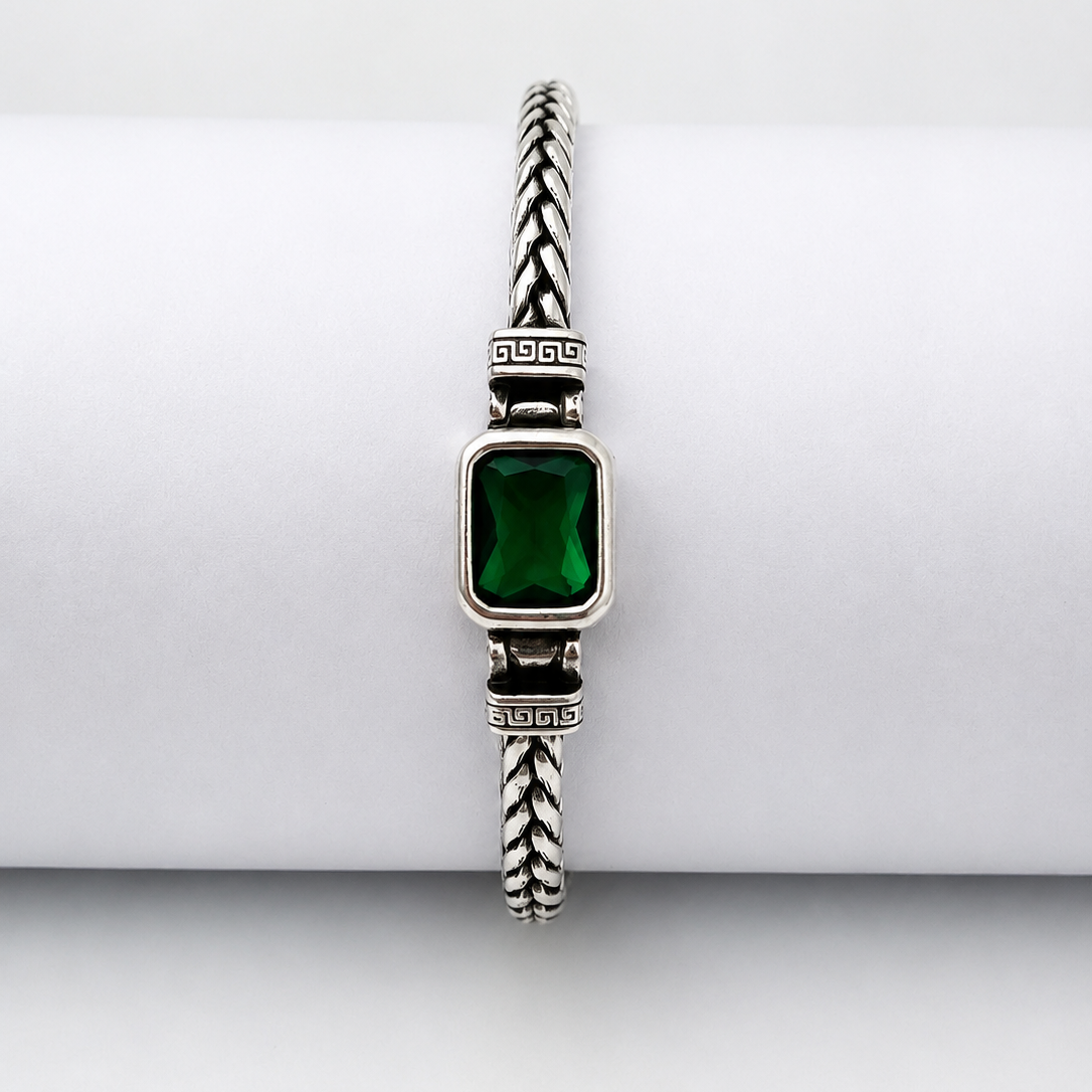 A silver-toned braided cable bracelet with a rectangular green gemstone centerpiece, displayed on a white cylindrical stand against a light gray background.