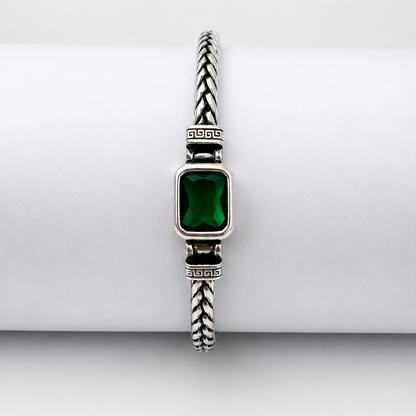 A silver-toned braided cable bracelet with a rectangular green gemstone centerpiece, displayed on a white cylindrical stand against a light gray background.
