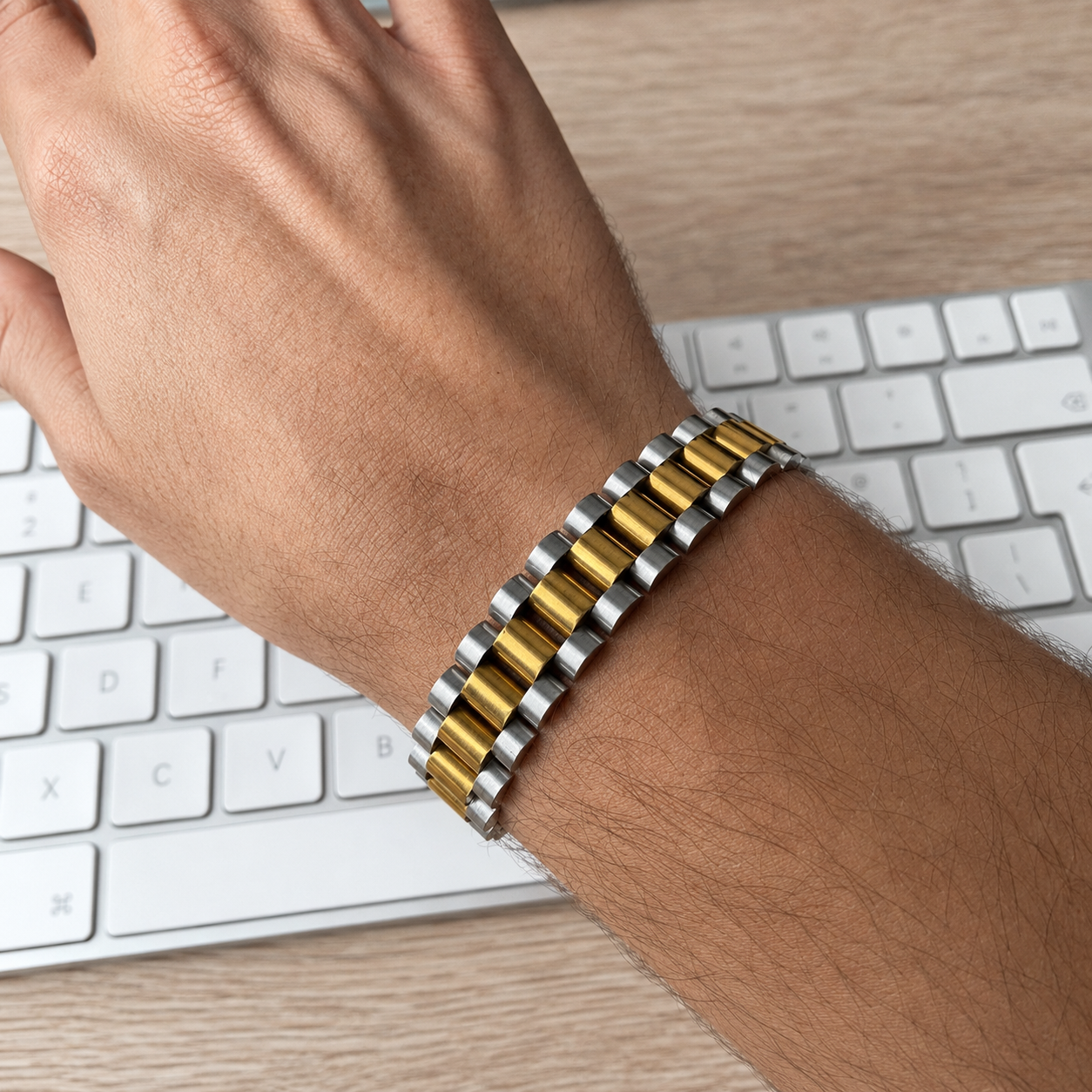 A close-up of a person's wrist wearing a two-tone metal link bracelet with alternating gold and silver cylindrical links, resting over a white keyboard on a light wood desk.