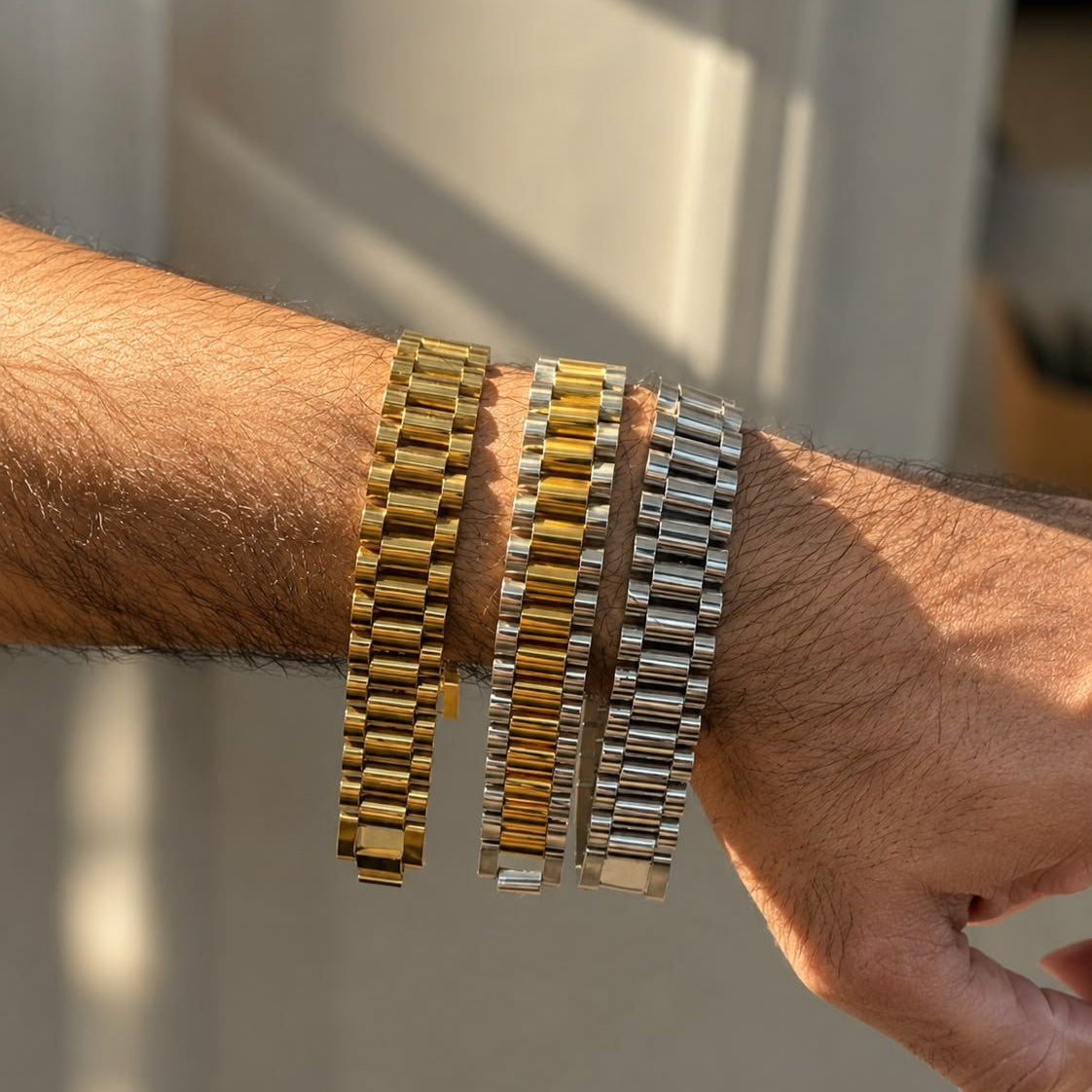 Three metal link bracelets in gold, two-tone gold and silver, and solid silver, displayed on a person's wrist against a softly blurred indoor background.