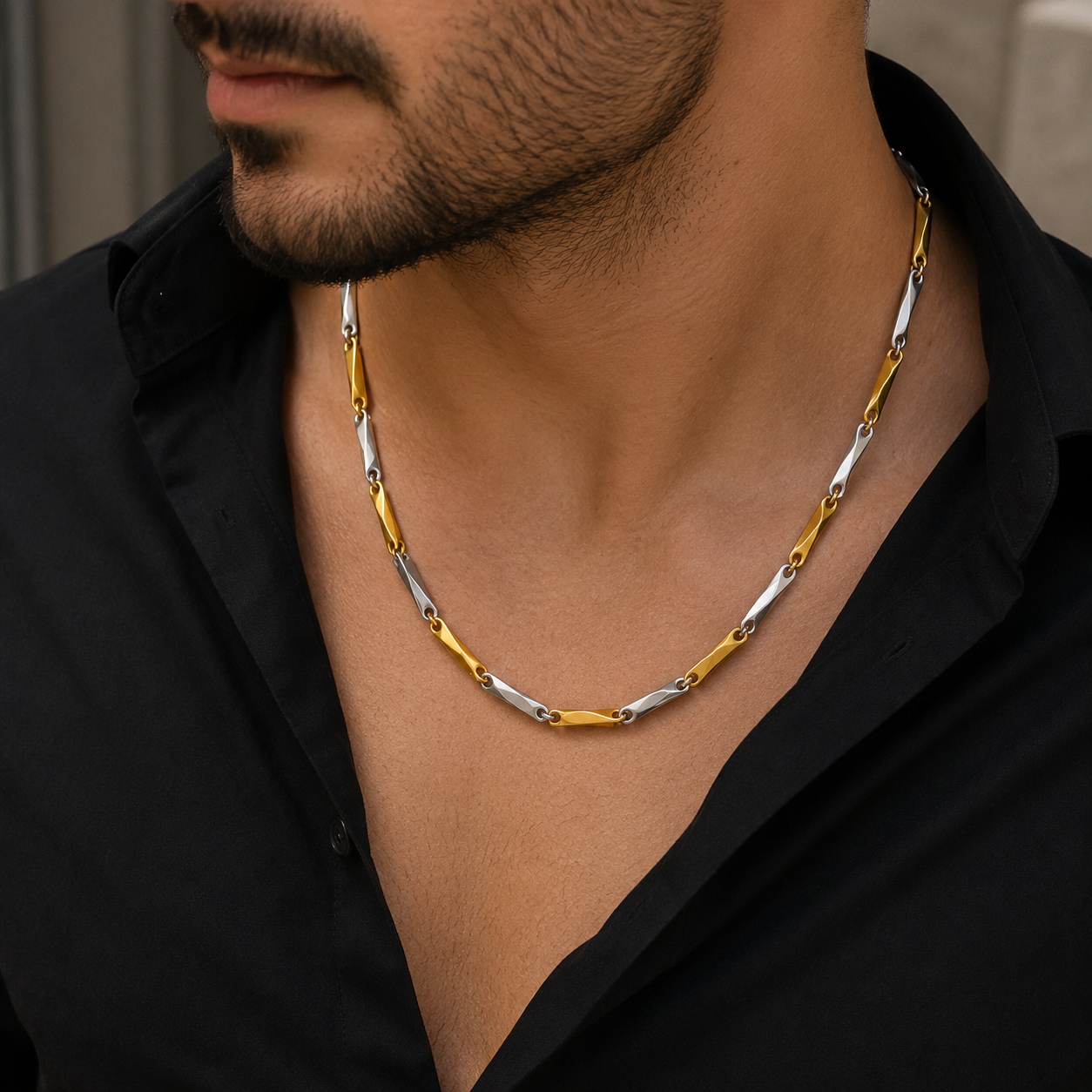 A close-up of a man wearing a two-tone gold and silver chain necklace with interlocking oval links, styled with a black button-down shirt.