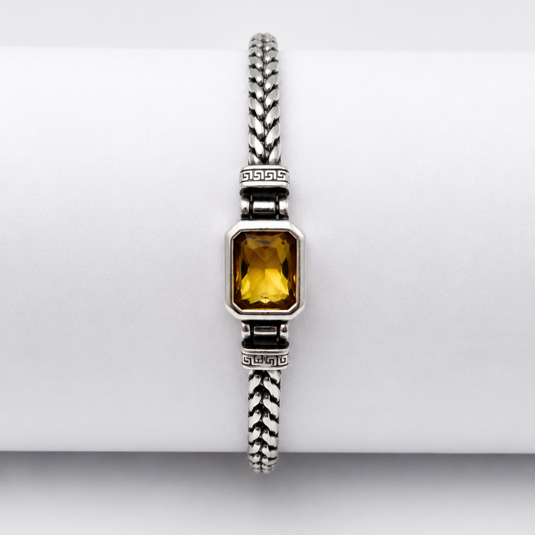 A silver-toned braided bracelet with a rectangular yellow gemstone, displayed on a white cylindrical stand.