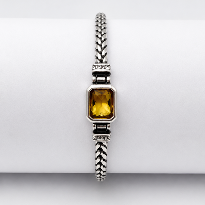 A silver-toned braided bracelet with a rectangular yellow gemstone, displayed on a white cylindrical stand.