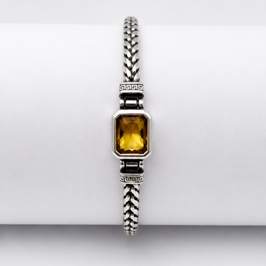 A silver-toned braided bracelet with a rectangular yellow gemstone, displayed on a white cylindrical stand.